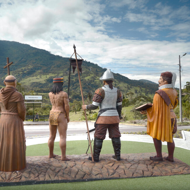 Vier lebensgroße Statuen von historischen Figuren stehen auf einem Podest im Freien vor einer Berglandschaft, darunter ein Mann in Rüstung, eine indigene Figur mit Kopfschmuck, ein Mönch mit Kreuz und ein Konquistator mit Buch und Umhang. // Four life-size statues of historical figures stand on a platform outdoors against a mountain landscape, including a man in armor, an indigenous figure with headgear, a monk with a cross, and a conquistador with a book and cloak.