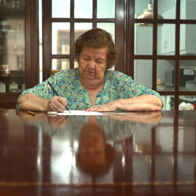 Ältere Frau sitzt an einem großen Holztisch und schreibt auf ein Blatt Papier, im Hintergrund ein Schrank mit Glasfront und eine Tür mit Glasfenstern. Older woman sitting at a large wooden table writing on a sheet of paper, with a glass-fronted cabinet and a door with glass panes in the background.