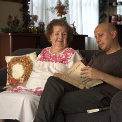 Zwei Personen sitzen auf einem dunkelgrauen Sofa, eine trägt ein weißes Kleid mit roten Stickereien, die andere ein schwarzes T-Shirt und hält ein Buch. // Two people sit on a dark gray sofa, one wearing a white dress with red embroidery, the other in a black t-shirt holding a book.