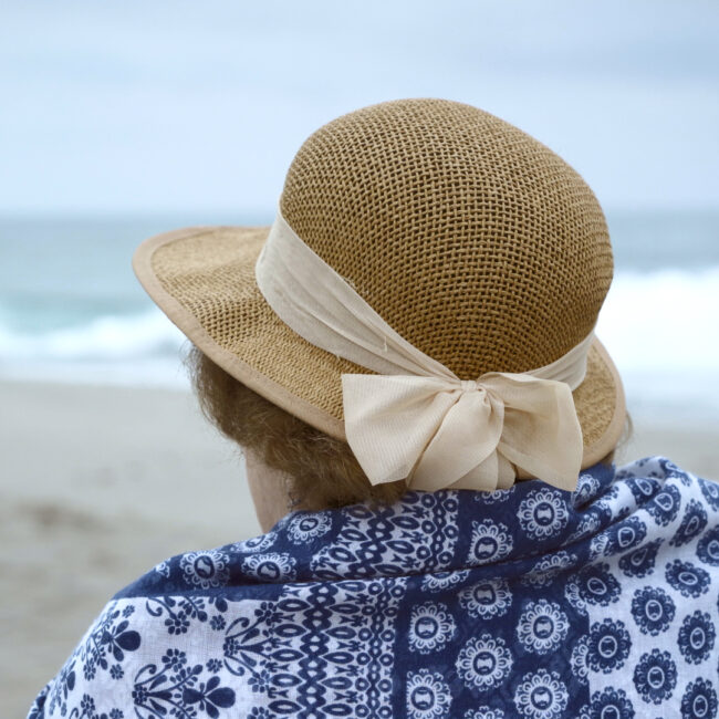 Ältere Frau mit strohfarbenem Hut mit weißem Band und blau-weiß gemustertem Tuch blickt aufs Meer am Strand. // Alderley woman wearing a straw hat with a white band and a blue-and-white patterned shawl looking at the sea on the beach.