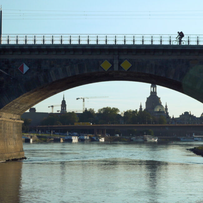 Brücke mit zwei Bögen über einen Fluss, Radfahrer auf dem Geländer, im Hintergrund Stadt mit historischen Gebäuden und Baukran. // Bridge with two arches over a river, cyclist on the railing, historic city buildings and a construction crane in the background.